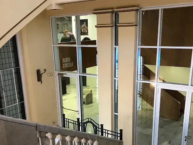 Vista aérea del interior de un edificio con puertas y ventanas de cristal. Una persona está sentada en un escritorio visible a través del cristal, y debajo hay una escalera.