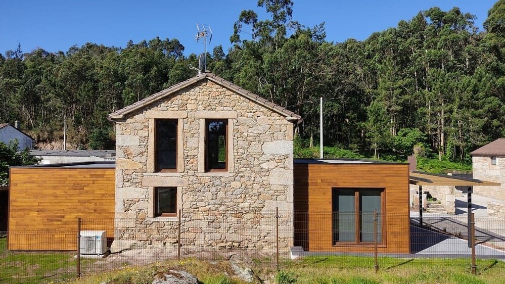 Una casa de piedra con una extensión de madera está rodeada de árboles.