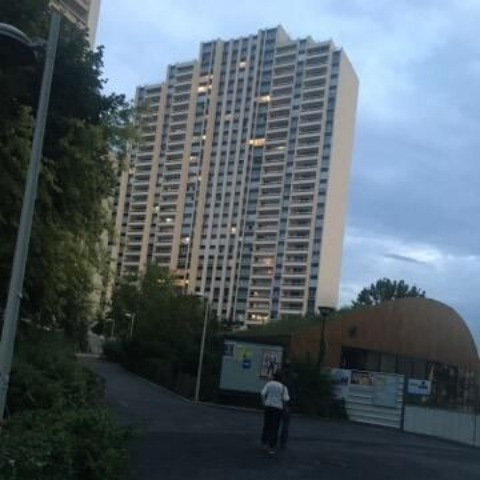 Immeuble d'appartements de grande hauteur sur une colline, avec une personne marchant sur la route devant. Ciel nuageux.