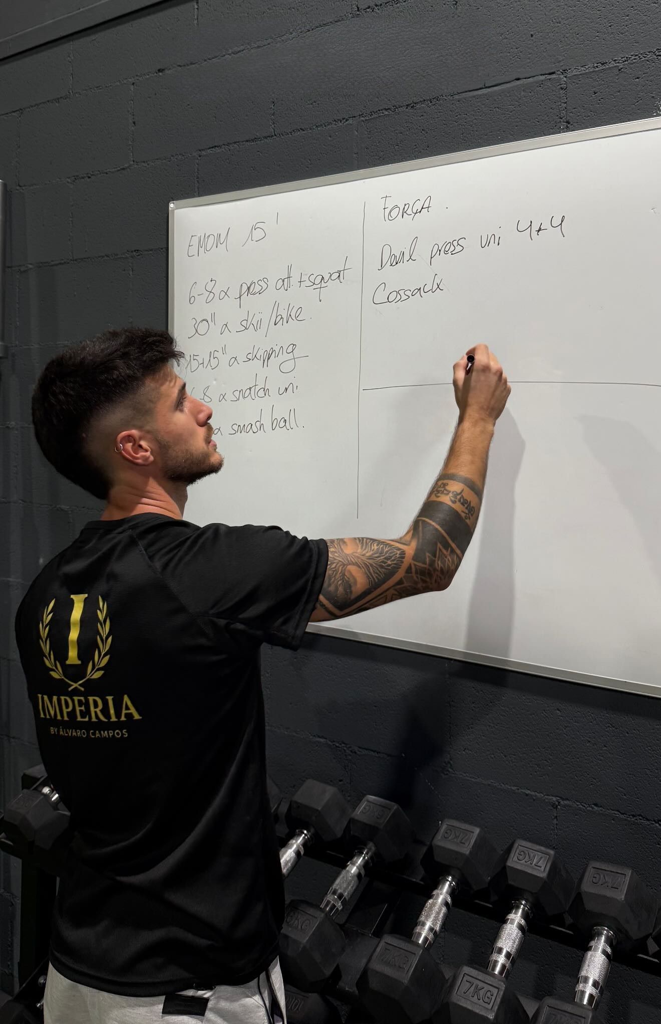 Una persona escribe en una pizarra blanca montada en la pared de un gimnasio, con mancuernas visibles debajo.