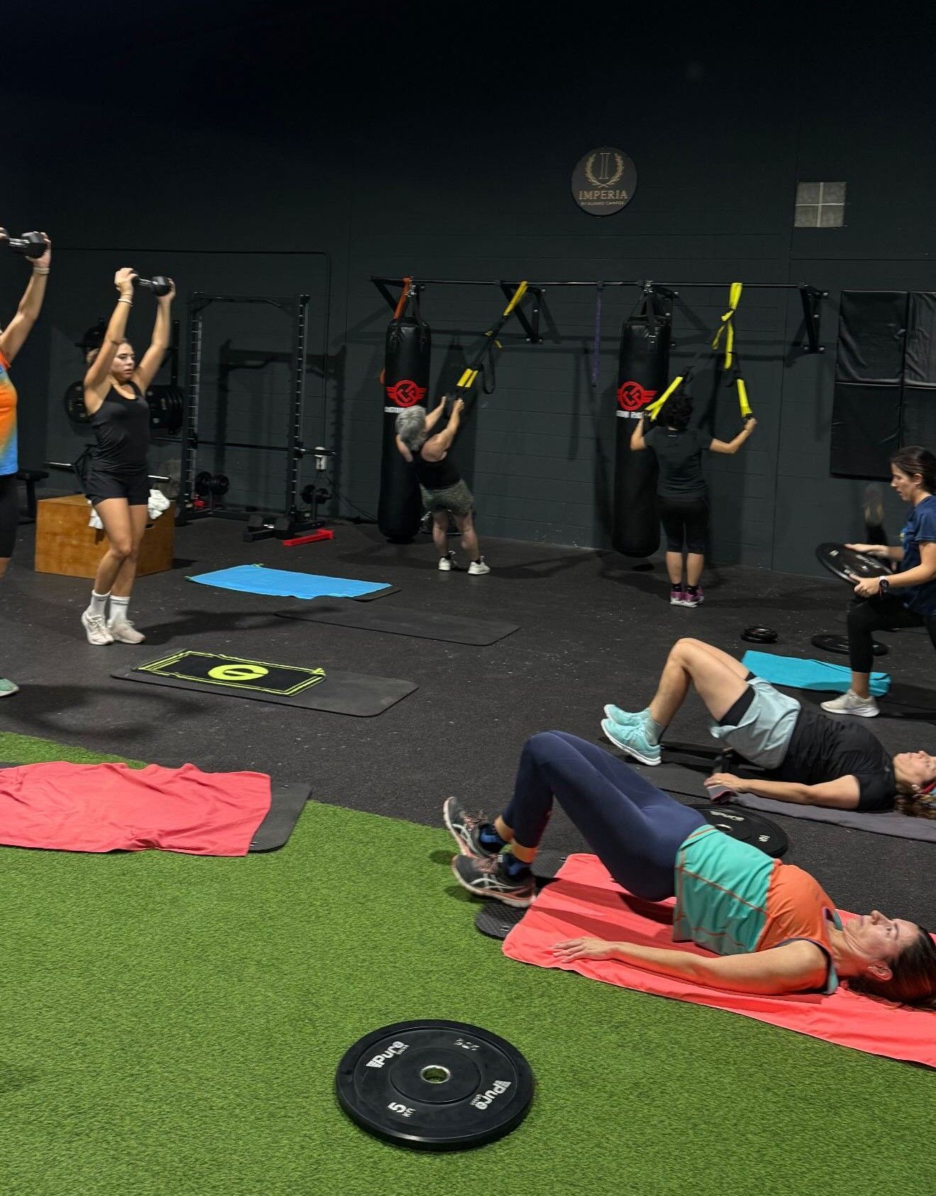 Una clase de fitness en un gimnasio donde la gente realiza varios ejercicios como elevaciones de cadera y press militar 