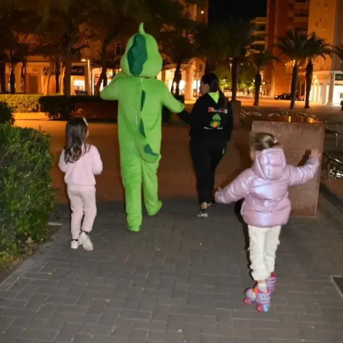 Niños con coloridos disfraces caminando de noche por una acera iluminada en un parque.