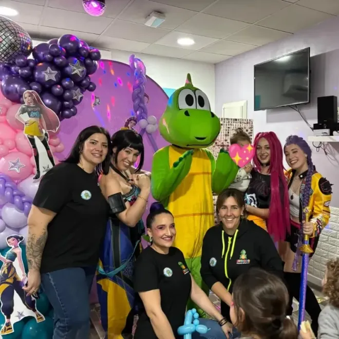 Grupo disfrazado posando en una celebración en interiores con globos y una mascota verde.