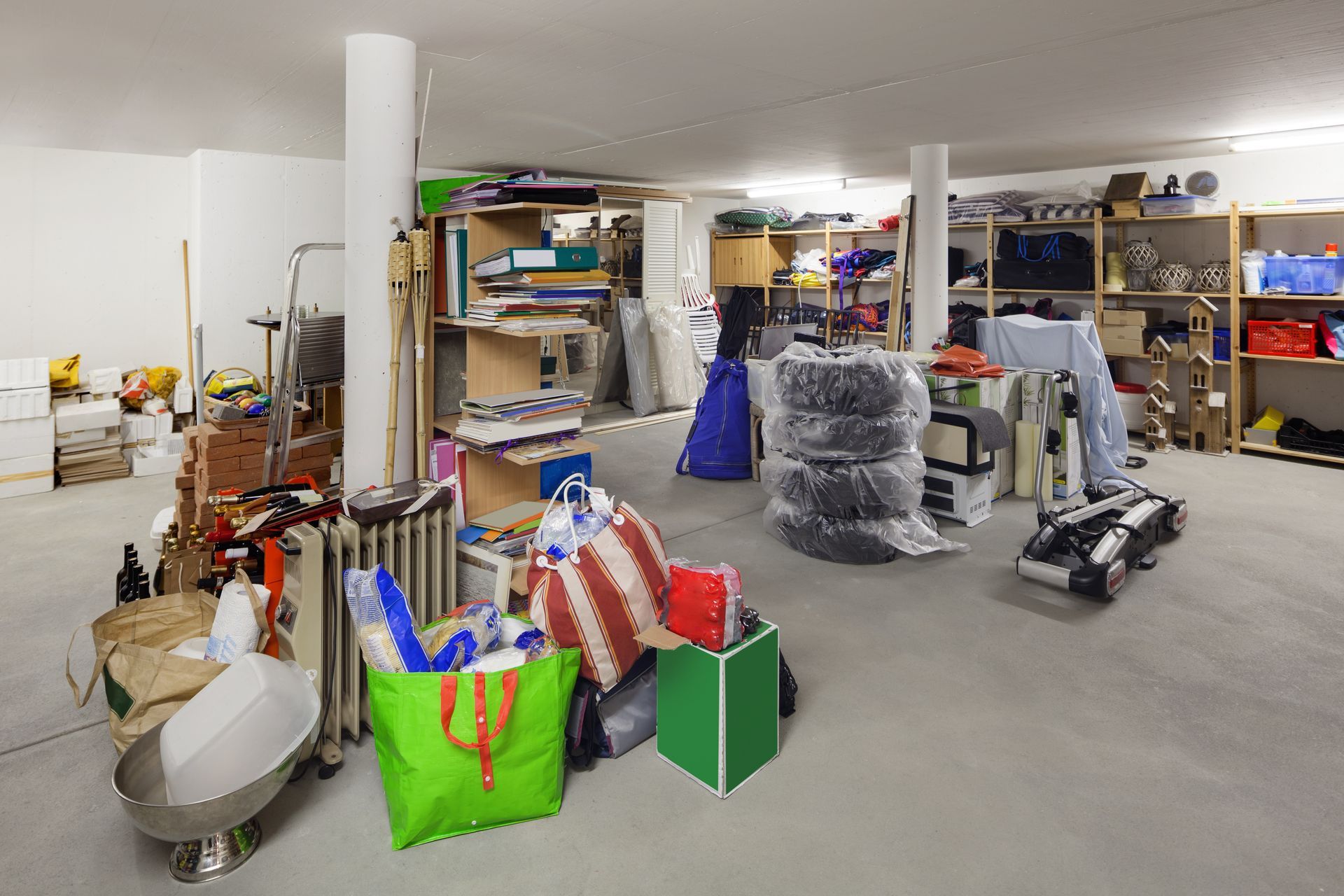Sous-sol encombré : pneus, étagères avec des boîtes, des sacs et divers objets contre des murs blancs et un sol en béton.