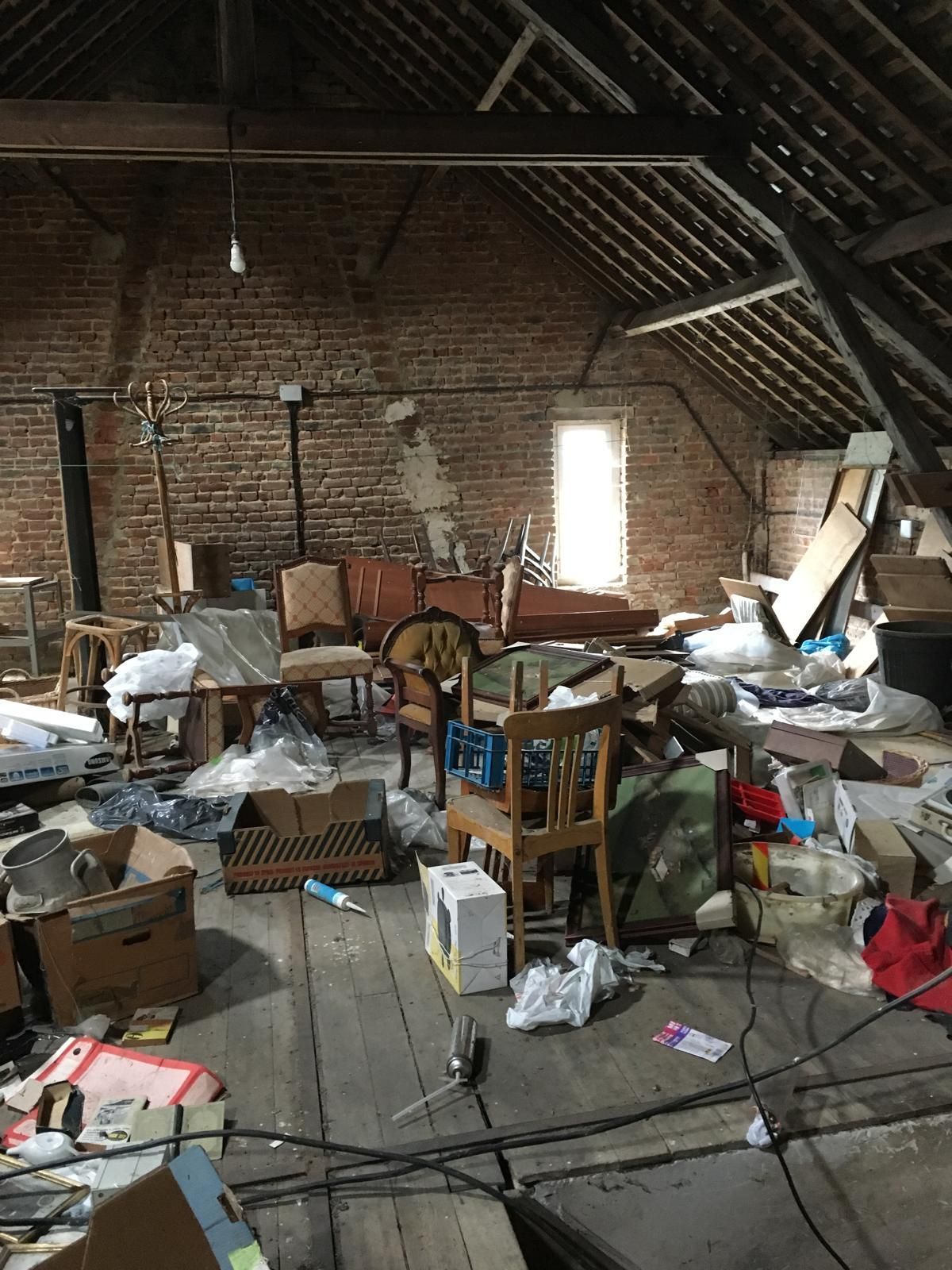 Grenier encombré avec plancher en bois, murs en briques apparentes et meubles et cartons divers.