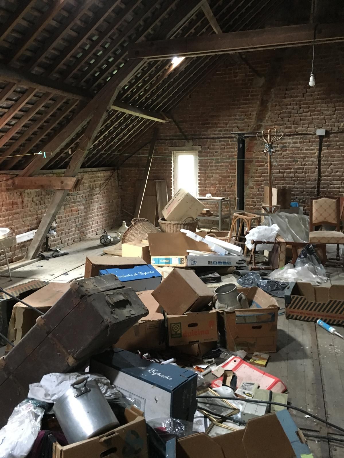 Un grenier en désordre rempli de cartons, des murs de briques, des poutres en bois et une petite fenêtre.