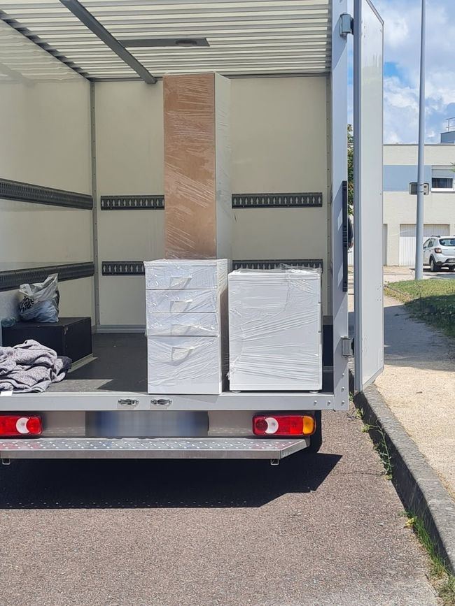 L'arrière ouvert d'un camion de déménagement stationné laisse apparaître deux meubles blancs emballés et un objet haut recouvert de carton.