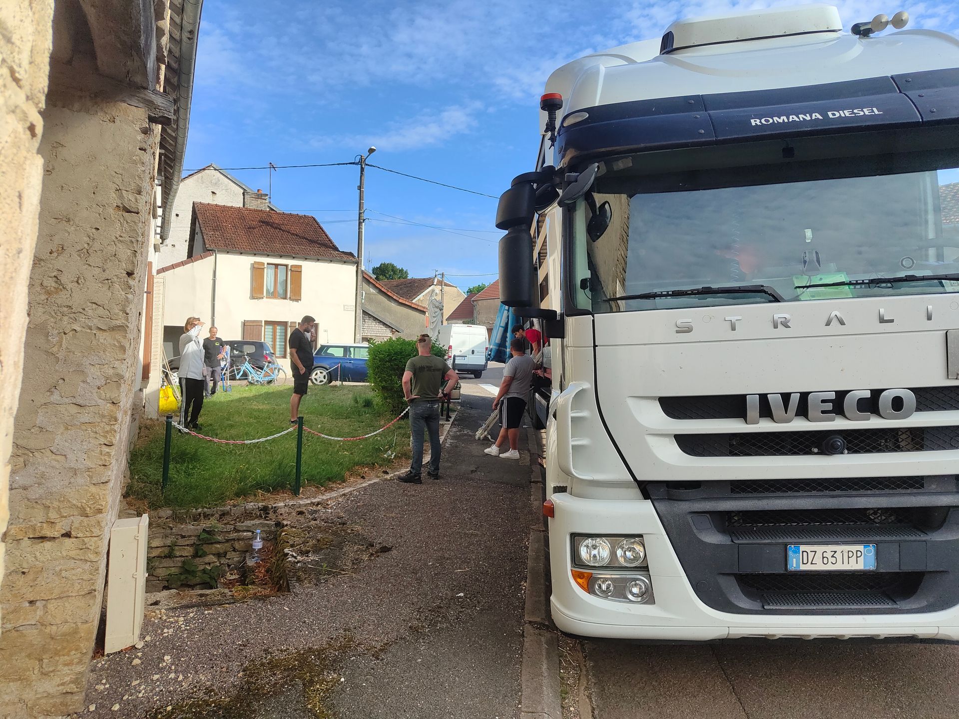 Un gros camion Iveco blanc est garé sur une route étroite, à côté d'un petit espace herbeux où se tiennent plusieurs personnes.
