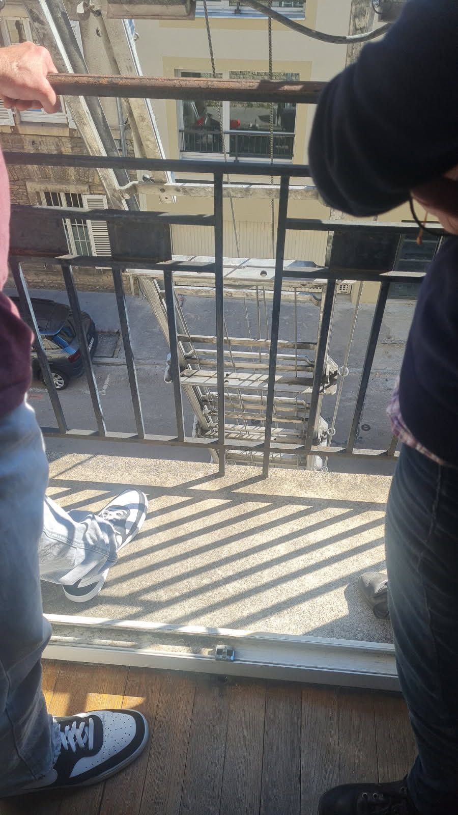 Deux personnes se tiennent sur un balcon donnant sur une rue, avec un escalier de secours métallique visible en contrebas.