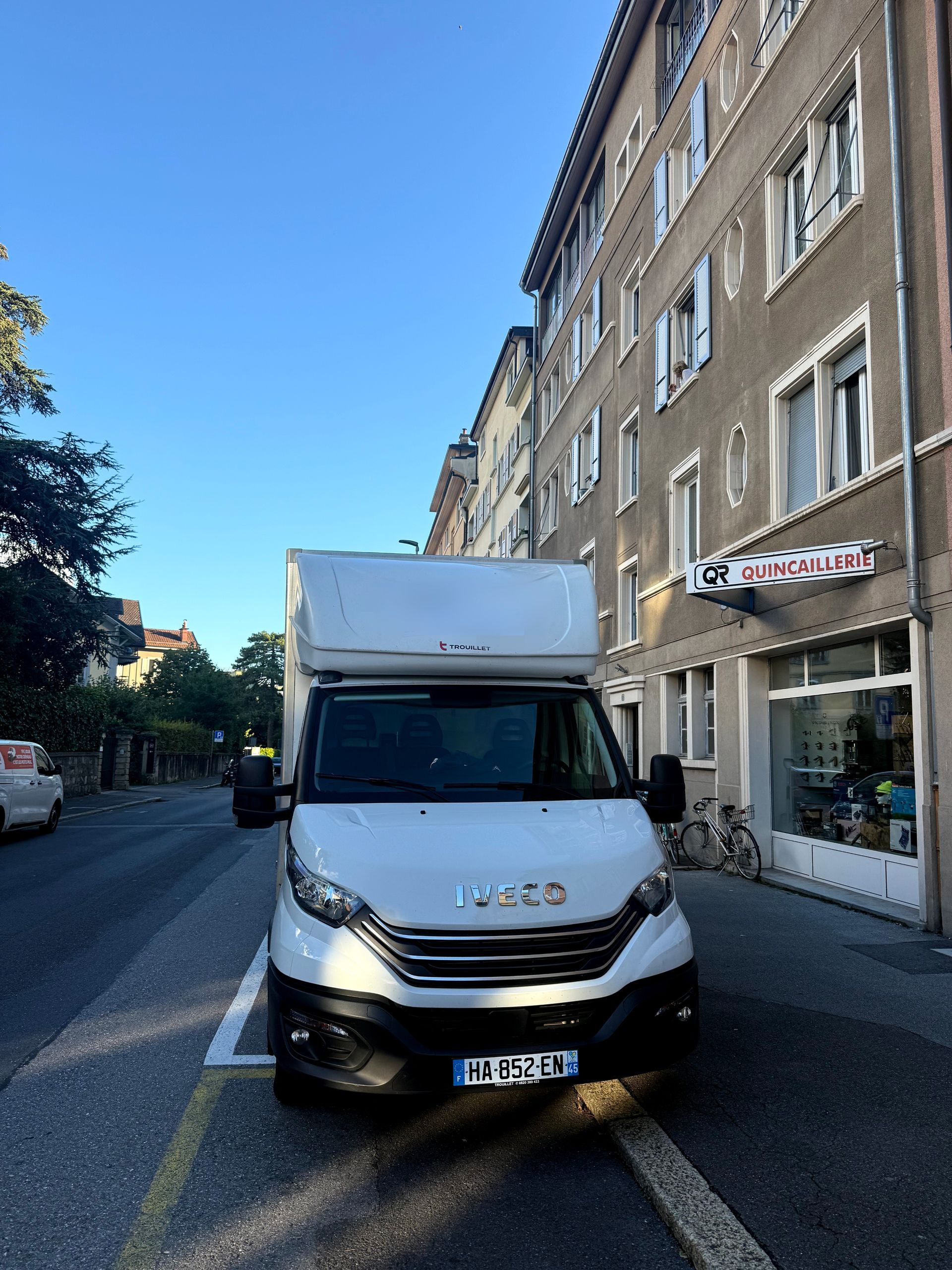 Un camion Iveco blanc est garé sur le côté d'une rue, à côté d'un immeuble de plusieurs étages abritant un magasin.