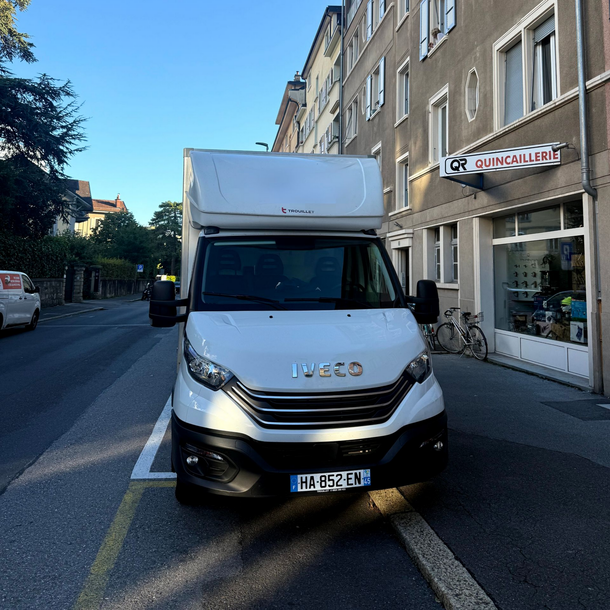 Un camion Iveco blanc est stationné dans une rue de la ville, devant un bâtiment portant une pancarte indiquant QUINCAILLERIE.