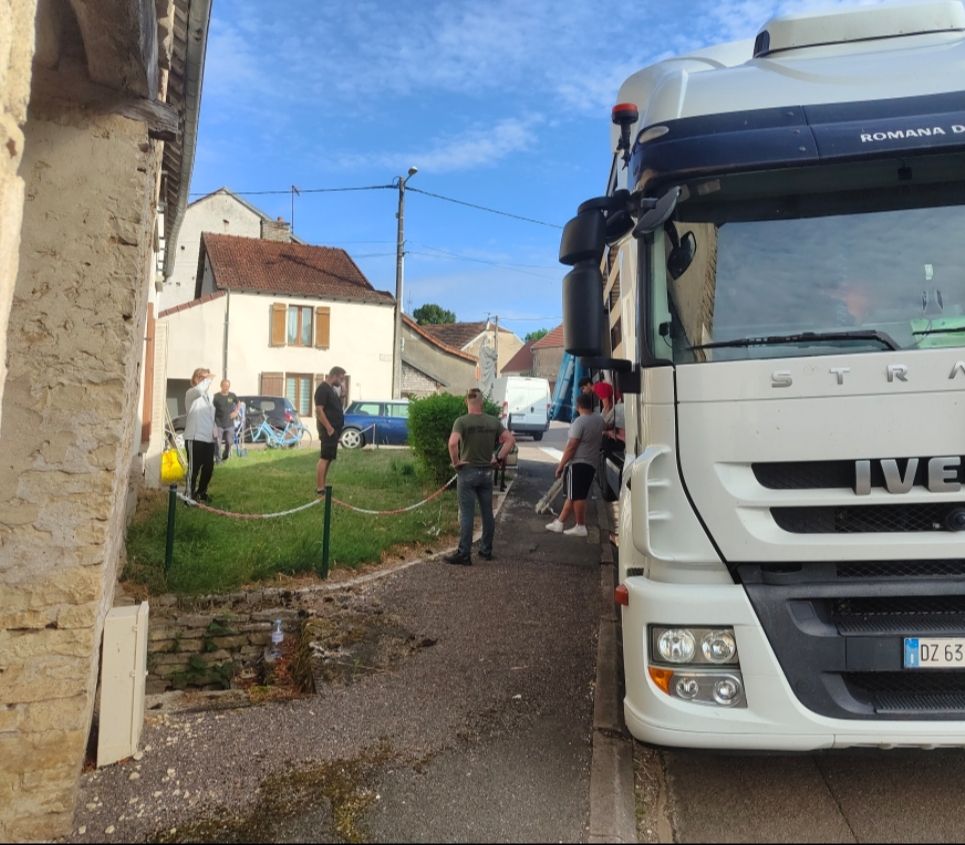 Un gros semi-remorque Iveco blanc est garé dans une rue étroite, à côté d'un espace vert où se tiennent plusieurs personnes.