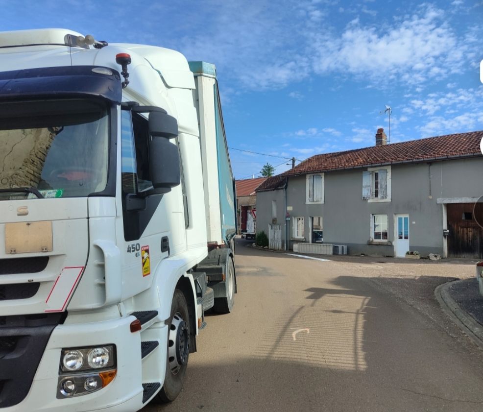 Un gros semi-remorque blanc est garé dans une rue étroite, à côté d'une maison aux couleurs claires, sous un ciel d'un bleu éclatant.