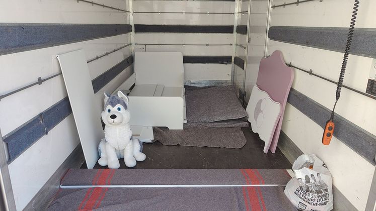 À l'intérieur d'un camion de déménagement, un husky en peluche est assis sur le sol à côté de meubles blancs démontés et d'une tête de lit rose.
