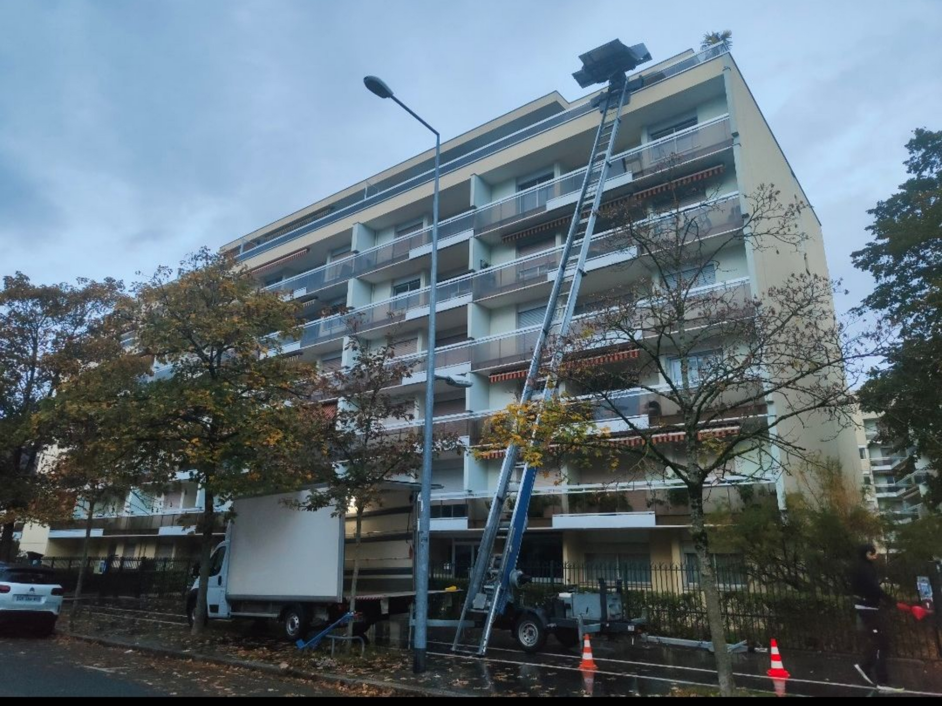 Un camion de déménagement blanc stationné dans la rue, avec un monte-meubles déployé jusqu'au balcon d'un immeuble d'appartements à plusieurs étages.