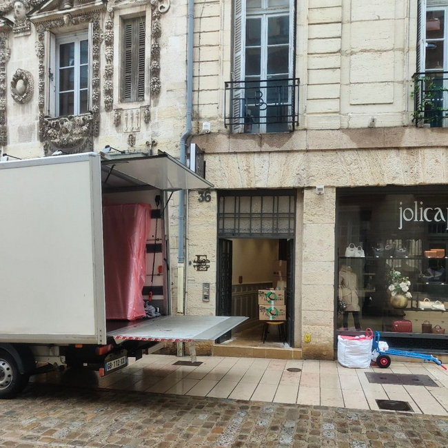 Un camion de livraison stationné dans une rue pavée, devant un bâtiment en pierre avec une entrée de magasin.