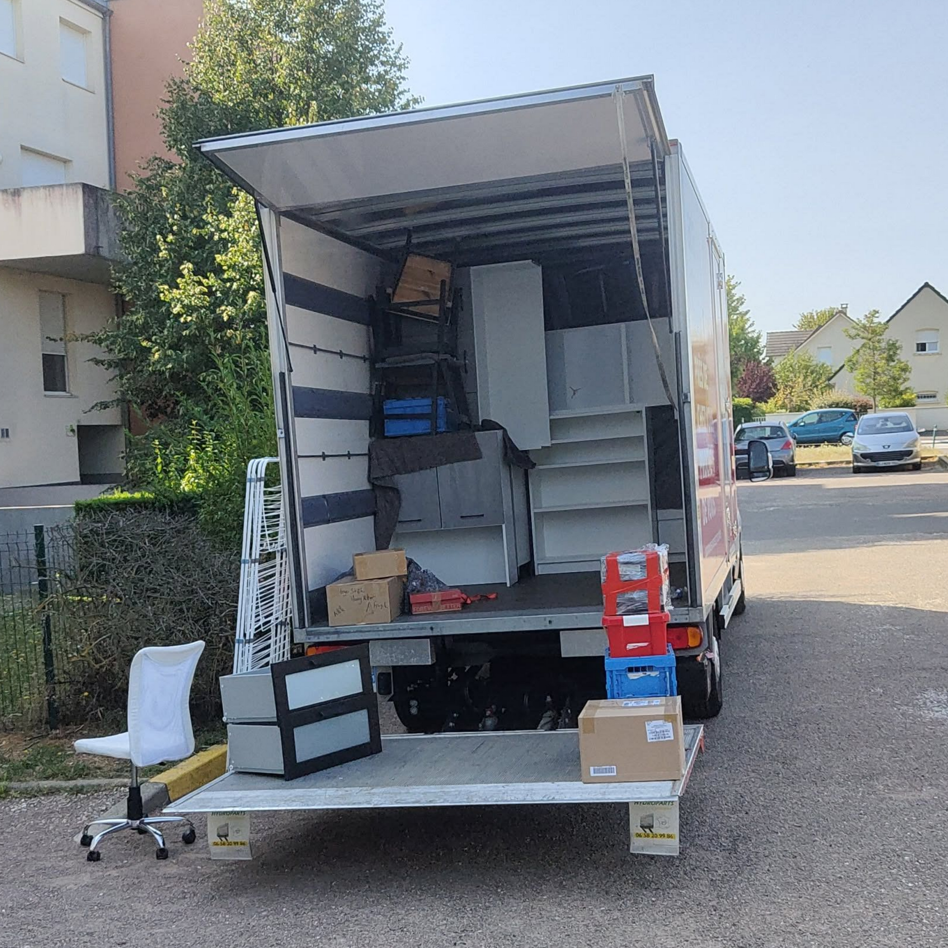 Un camion de déménagement est garé à l'extérieur, sa benne ouverte remplie de meubles et de cartons, et une chaise de bureau blanche se trouve à proximité.