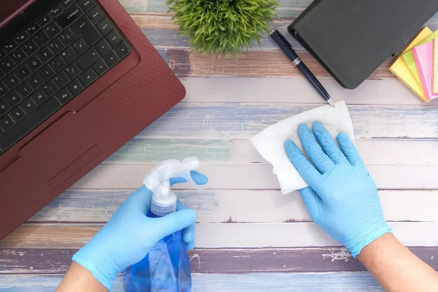 Una persona que lleva guantes azules está limpiando un escritorio con una botella de spray.