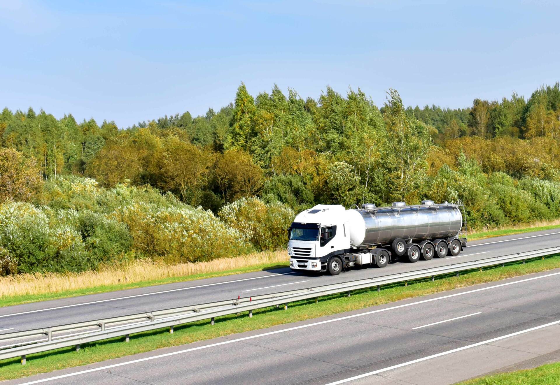 Valkoinen säiliöauto ajaa moottoritietä rehevän, vihreän metsän vieressä aurinkoisena päivänä.