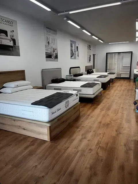 Interior de tienda de colchones con varias camas en exposición, suelo de madera y carteles en paredes blancas.