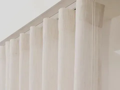 Cortinas blancas transparentes que cuelgan de un riel en el techo y crean pliegues verticales contra una pared blanca.