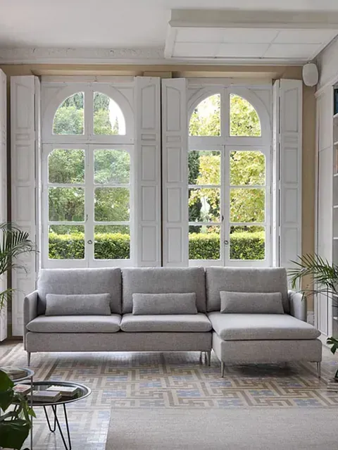Sofá seccional gris en una habitación luminosa con grandes ventanas arqueadas y follaje verde en el exterior.