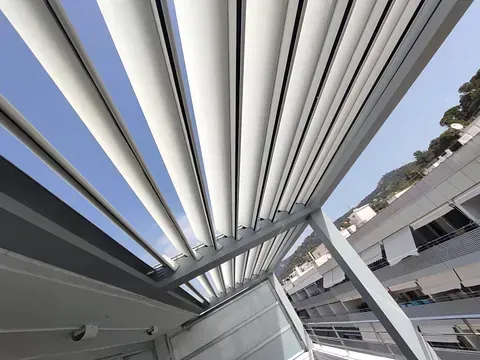 Vista de listones de techo con lamas de aluminio contra un cielo azul brillante, sombreando parcialmente un área al aire libre.