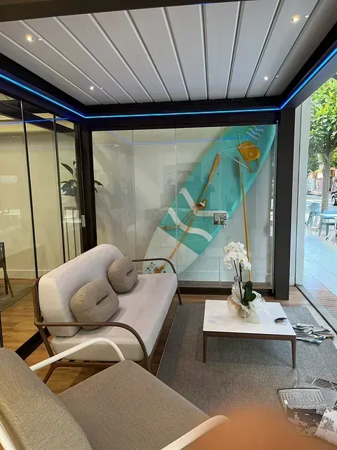 Un interior de sala de exposición lleno de luz, con una tabla de surf, asientos y un ambiente interior-exterior.