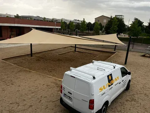Una camioneta SUNBAY blanca estacionada en un arenero bajo velas de sombra en un parque.