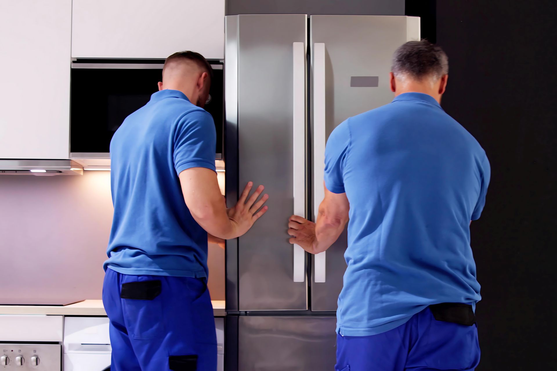 Deux déménageurs en uniforme bleu transportent un réfrigérateur en acier inoxydable dans une cuisine