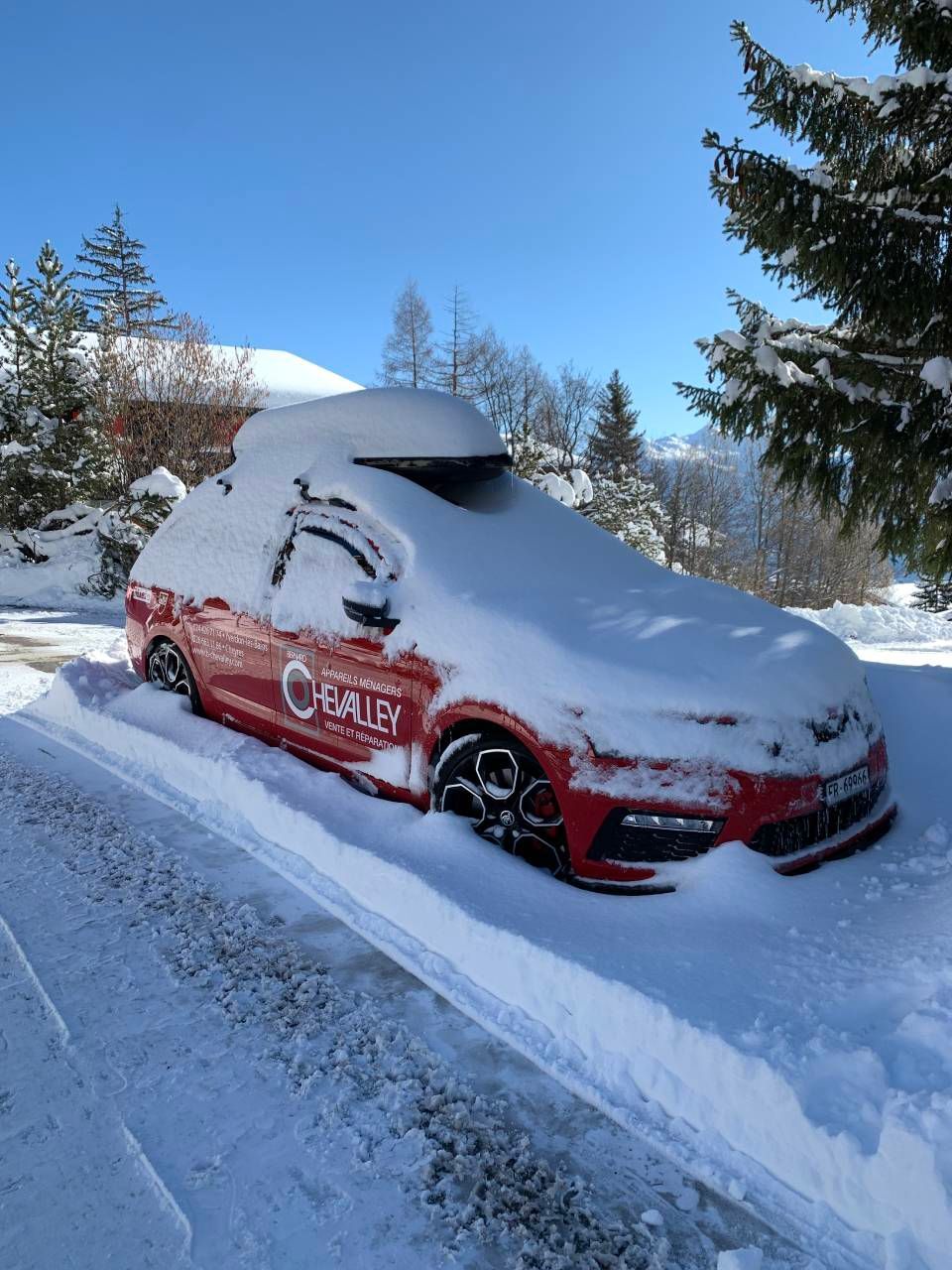 Voiture rouge ensevelie sous la neige au bord d'une route enneigée, sous un ciel bleu azur.