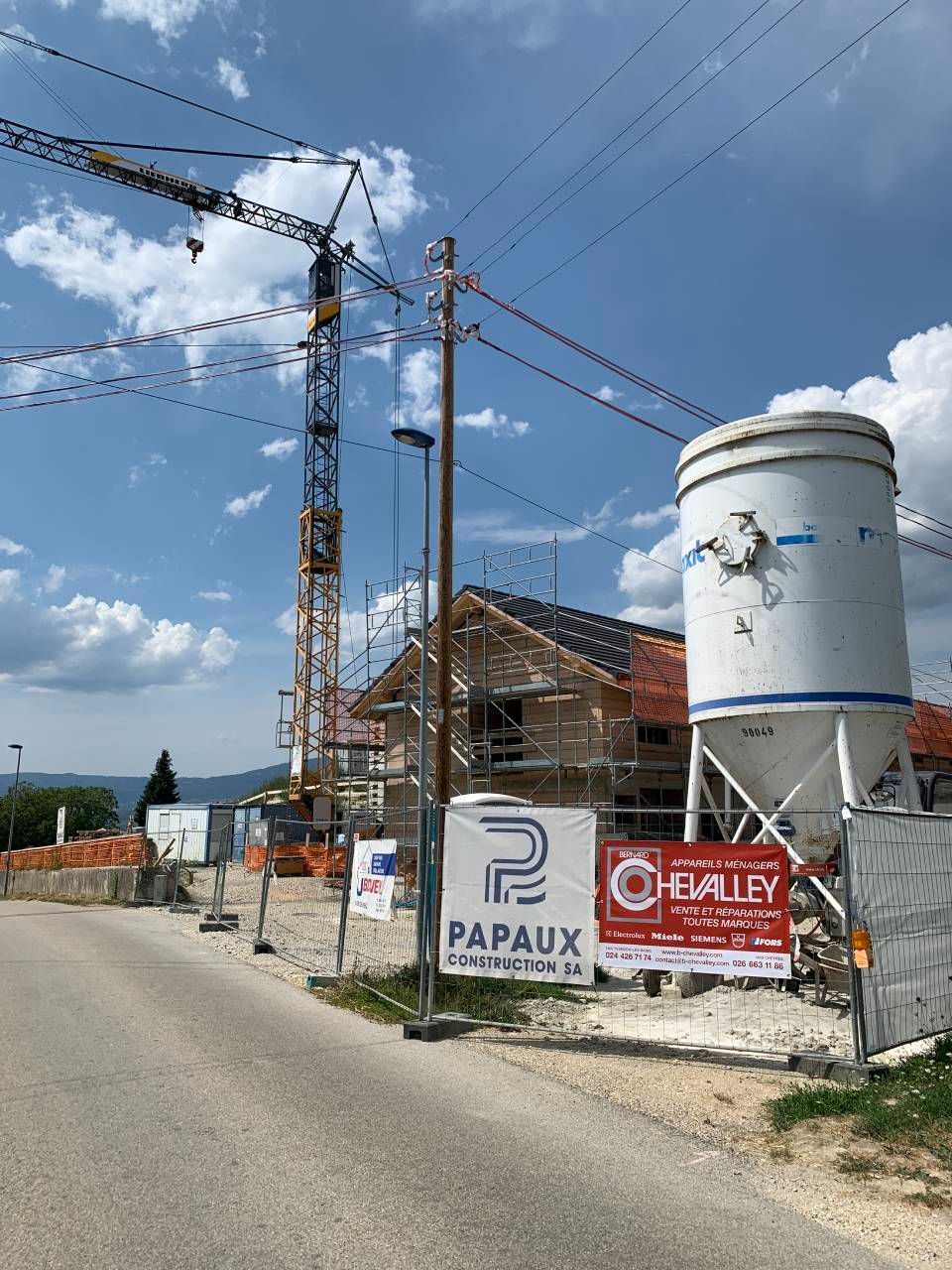 Chantier routier avec des grues, un silo blanc et des panneaux sous un ciel bleu.