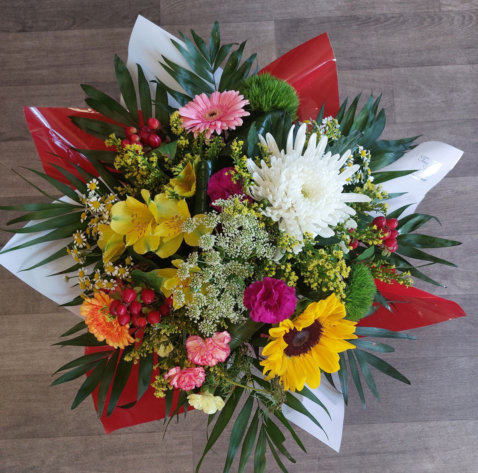 Bouquet de fleurs colorées, comprenant un tournesol, un gerbera et un chrysanthème, enveloppé dans du papier rouge et blanc.