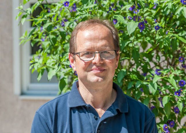 Mann in blauem Hemd steht im Freien und lächelt vor grünem Laub und lila Blüten.