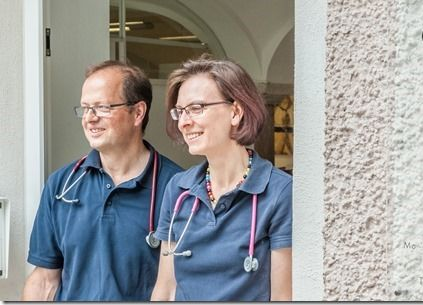 Zwei lächelnde Menschen stehen an einer Steinmauer mit einer Namenstafel.