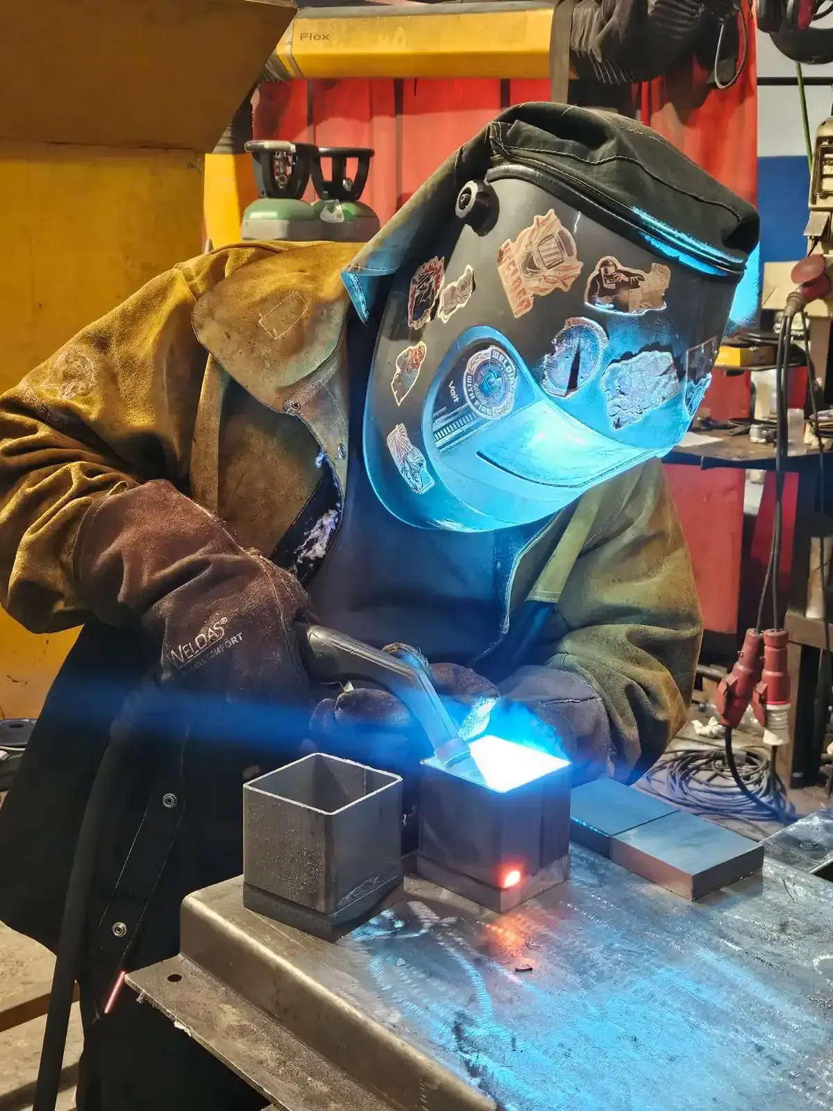 Soldador con traje protector azul soldando una tubería de metal, saltan chispas en un taller industrial.