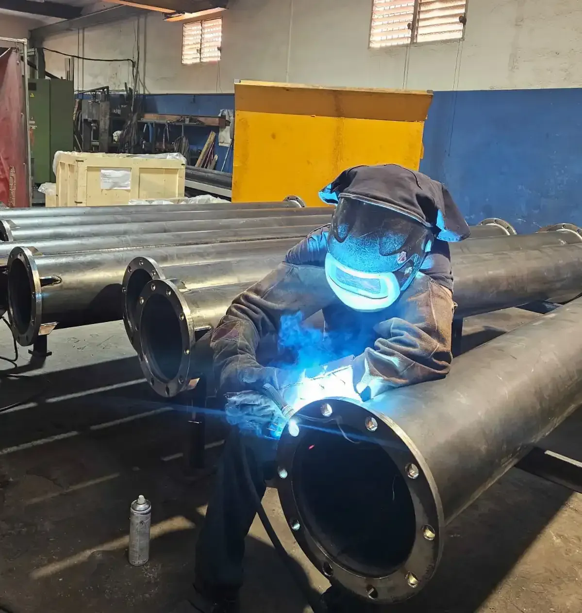 Soldador con traje de protección azul soldando una tubería de metal con chispas brillantes en un taller industrial.