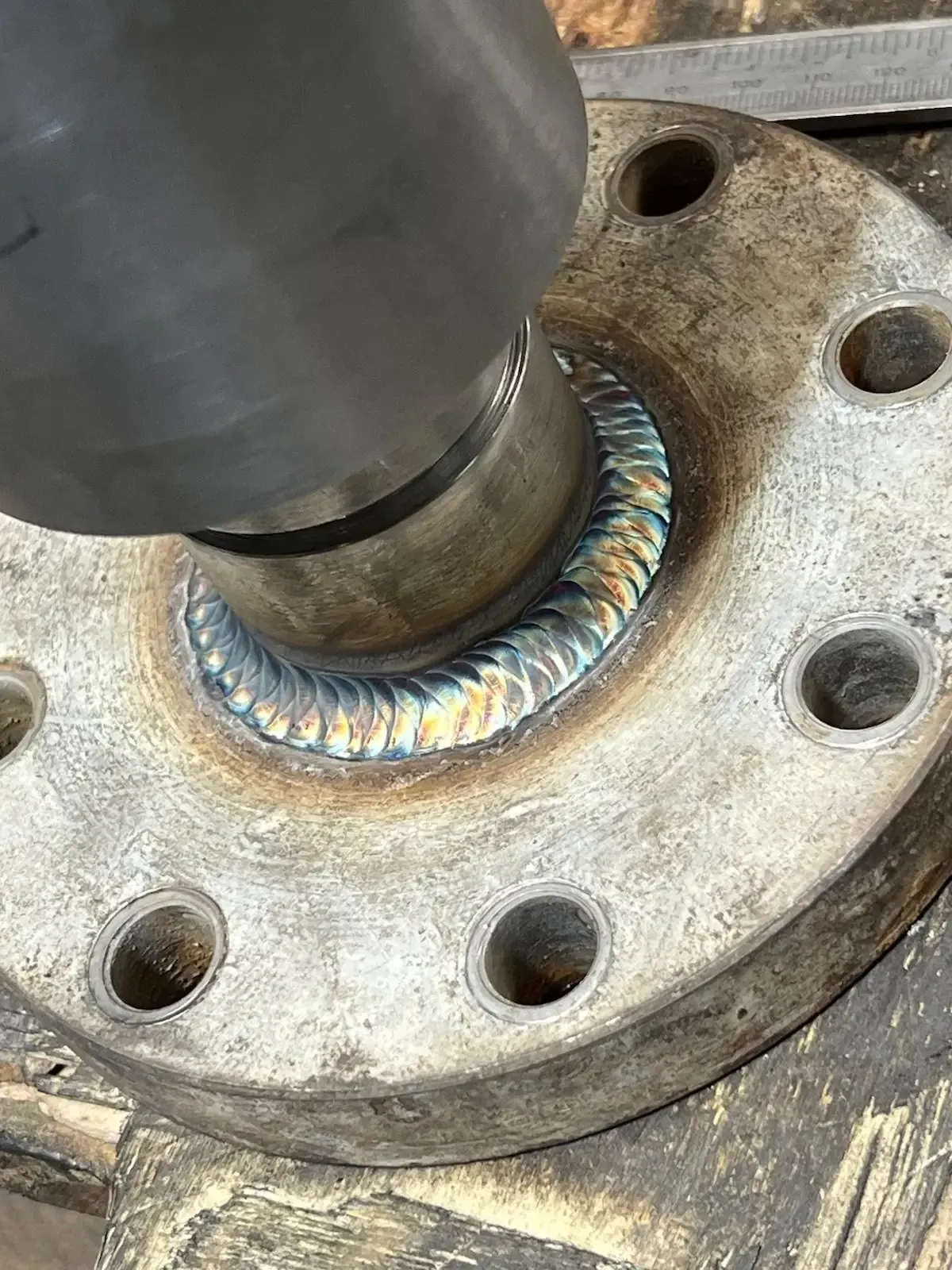 Primer plano de una brida metálica soldada con orificios para pernos y un cordón de soldadura de colores
