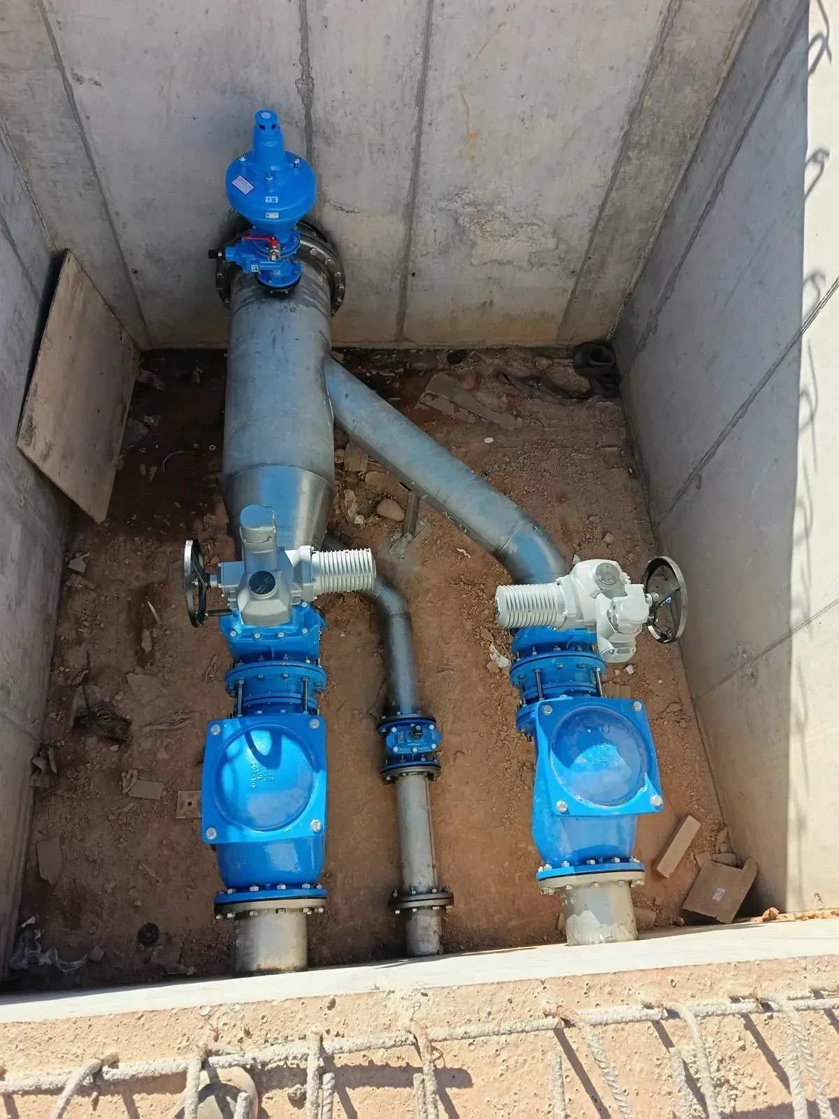 Tuberías y válvulas subterráneas de agua de color azul en una fosa de hormigón, con escombros en el fondo.