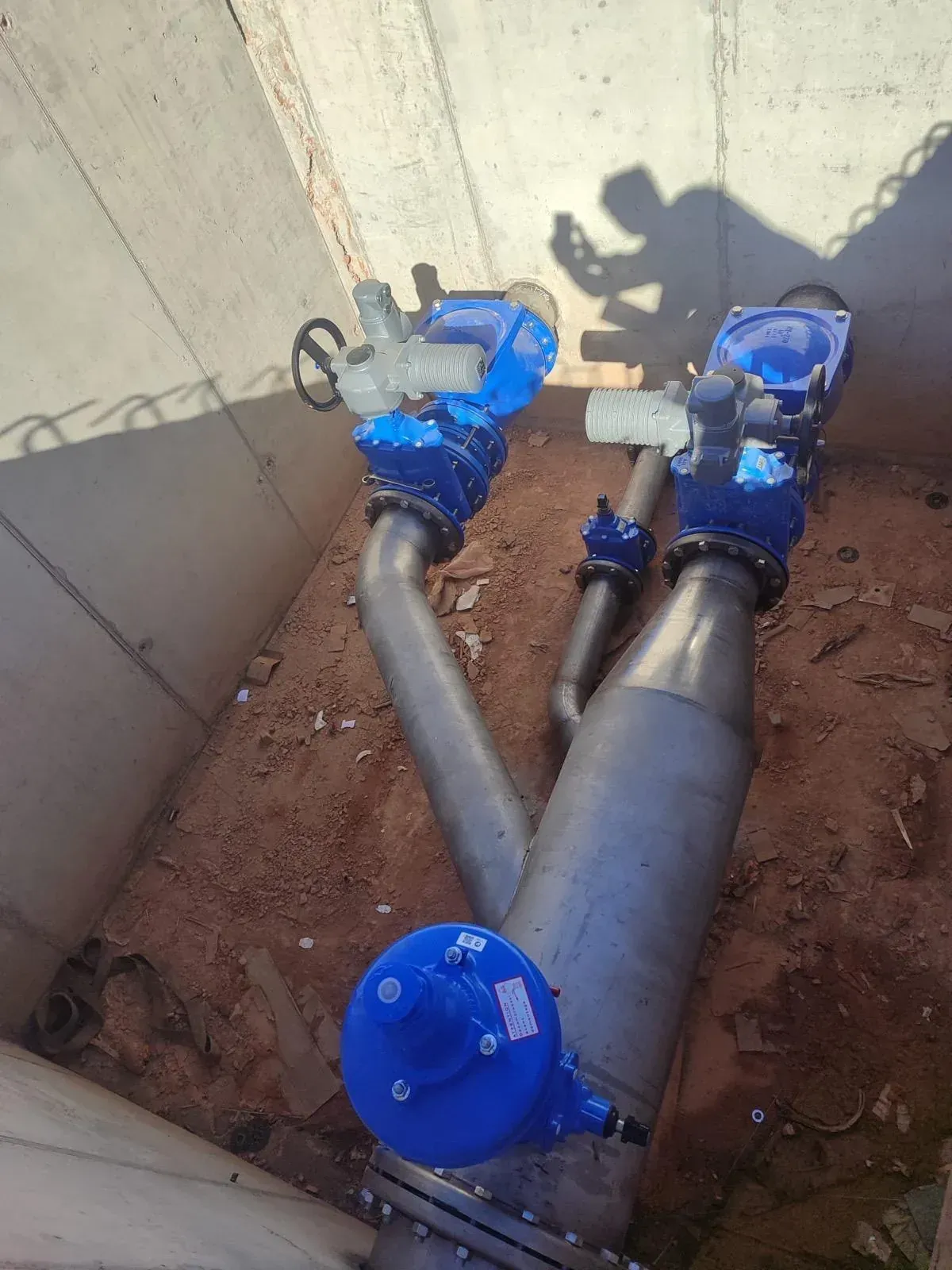 Tuberías y válvulas industriales de color azul en una zanja estrecha junto a un muro de hormigón.