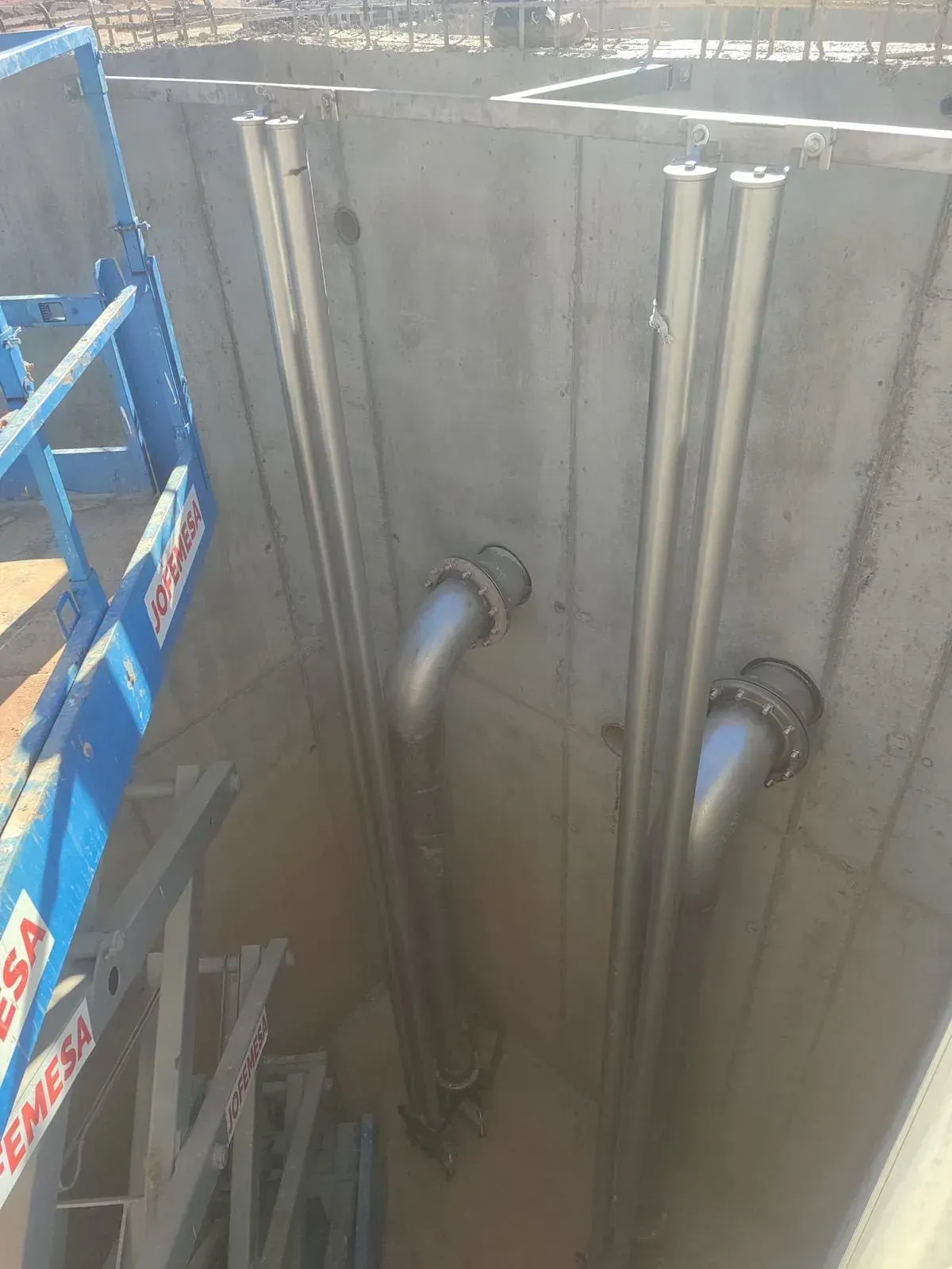 Zanja de obra con paredes de hormigón, tuberías expuestas y andamios azules a la izquierda.