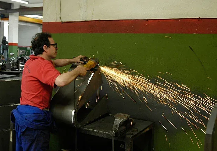 Un trabajador con camisa roja usa una amoladora, lanzando chispas a través de la pared del taller.