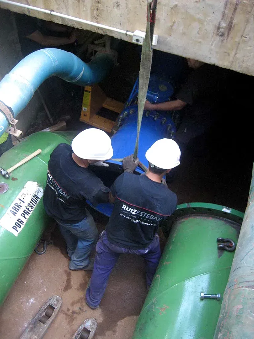 Obreros con cascos blancos inspeccionan una zanja húmeda con grandes tuberías verdes debajo de una estructura de hormigón.