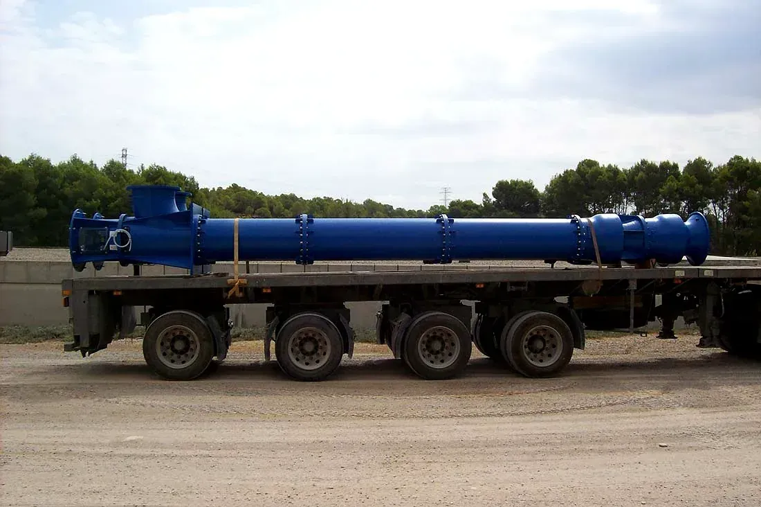 Camión cisterna azul sobre un remolque de plataforma, estacionado sobre grava con árboles al fondo.