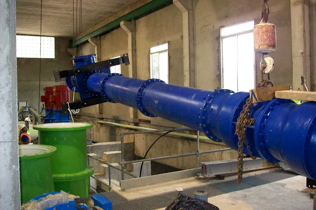 Una gran tubería industrial azul se encuentra dentro de un cuarto de servicio de hormigón con barriles verdes cerca.