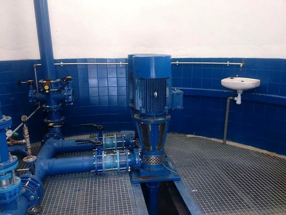 Bombas y tuberías industriales azules en un cuarto de servicio alicatado con suelo de rejilla metálica y fregadero de pared.