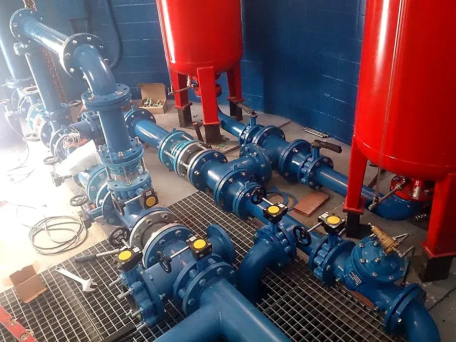 Tuberías industriales azules y depósitos rojos en una sala de máquinas, con válvulas y rejillas en el suelo.