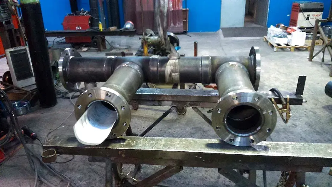 Conjunto de tubo de escape metálico sobre un banco de trabajo en un garaje o taller, con tubos y bridas plateadas.