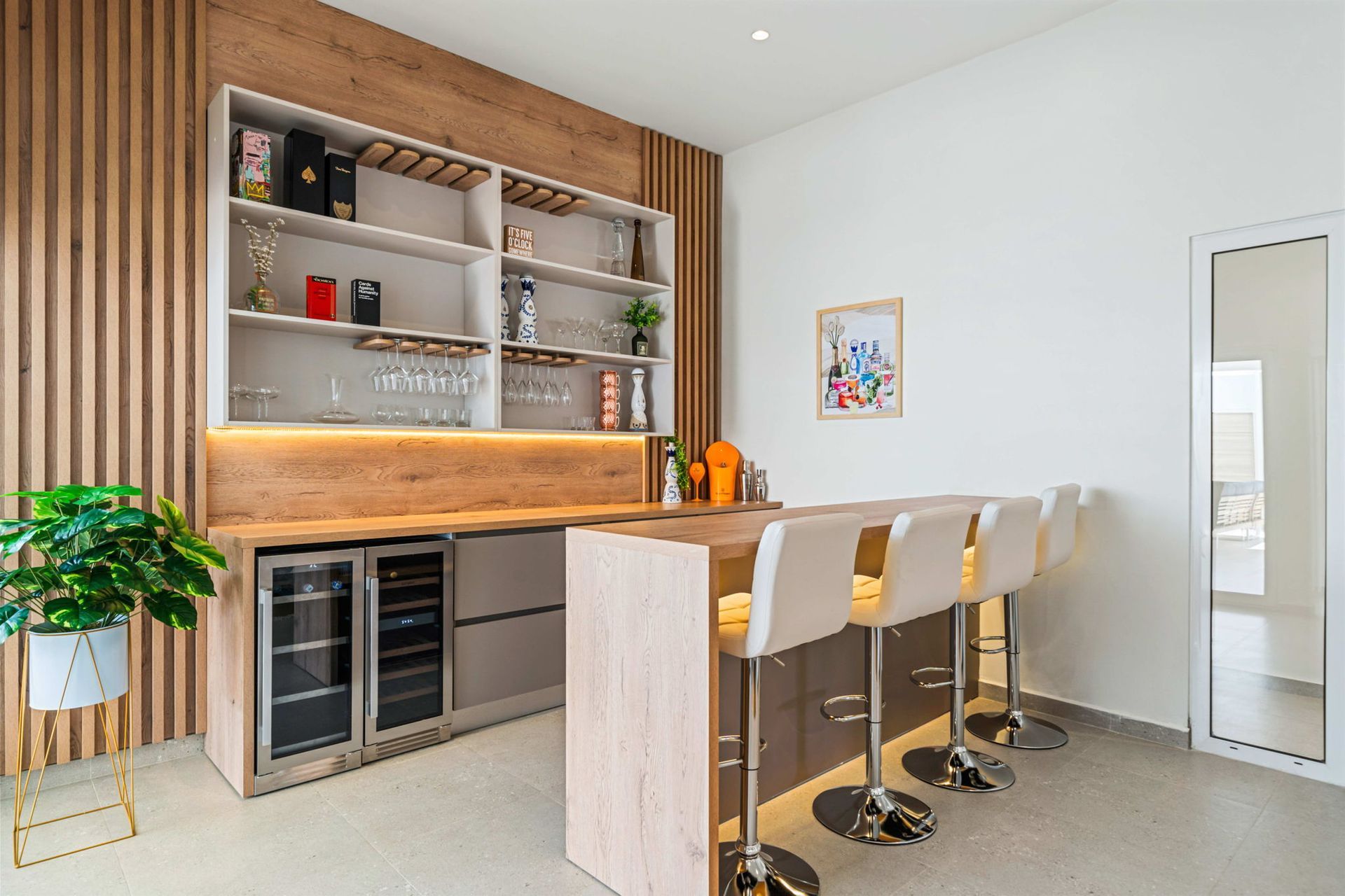 Modern home bar with wooden counter, open shelves, and four white stools in a bright room