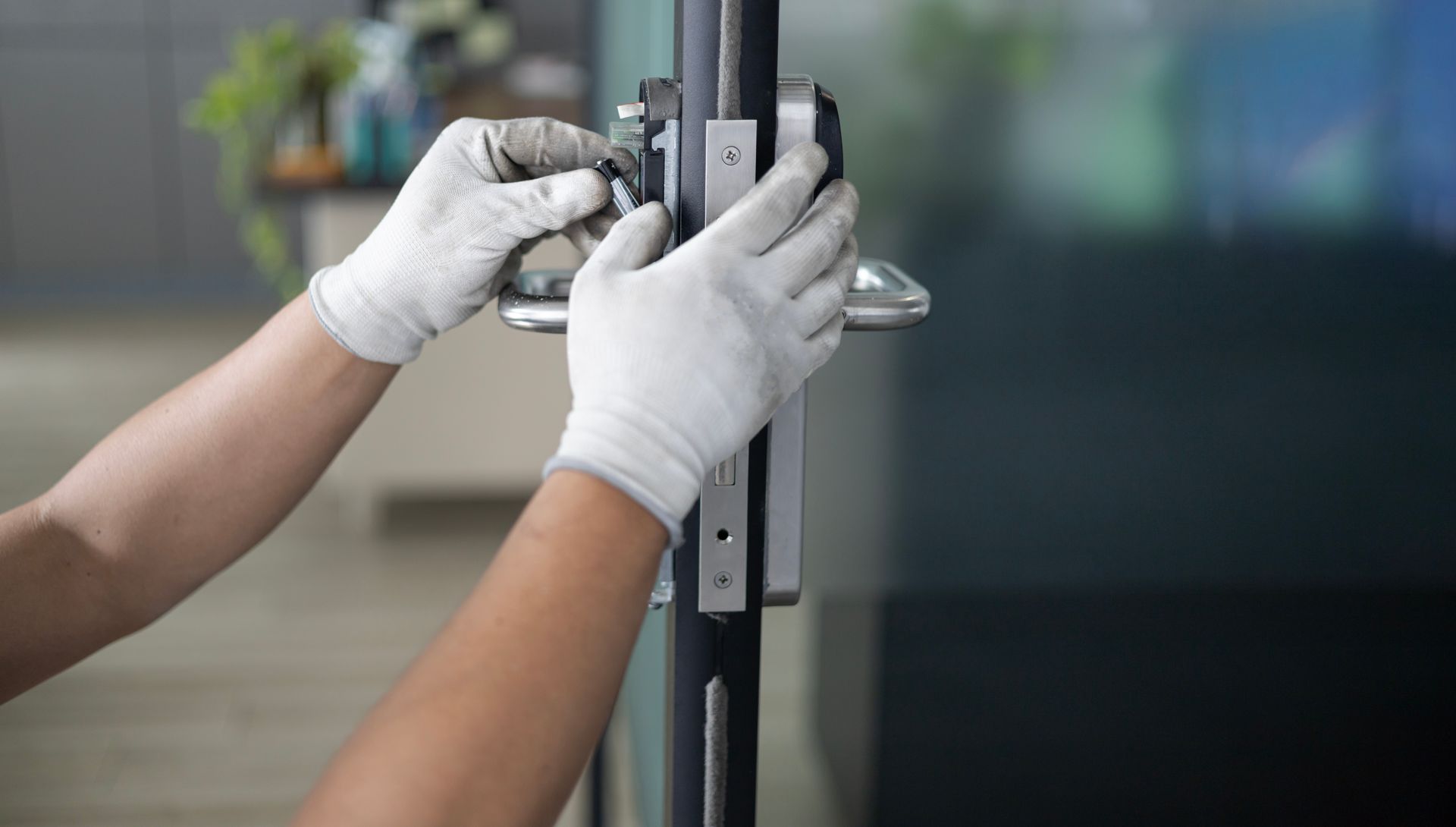Des mains gantées de blanc installent une serrure sur une porte vitrée.